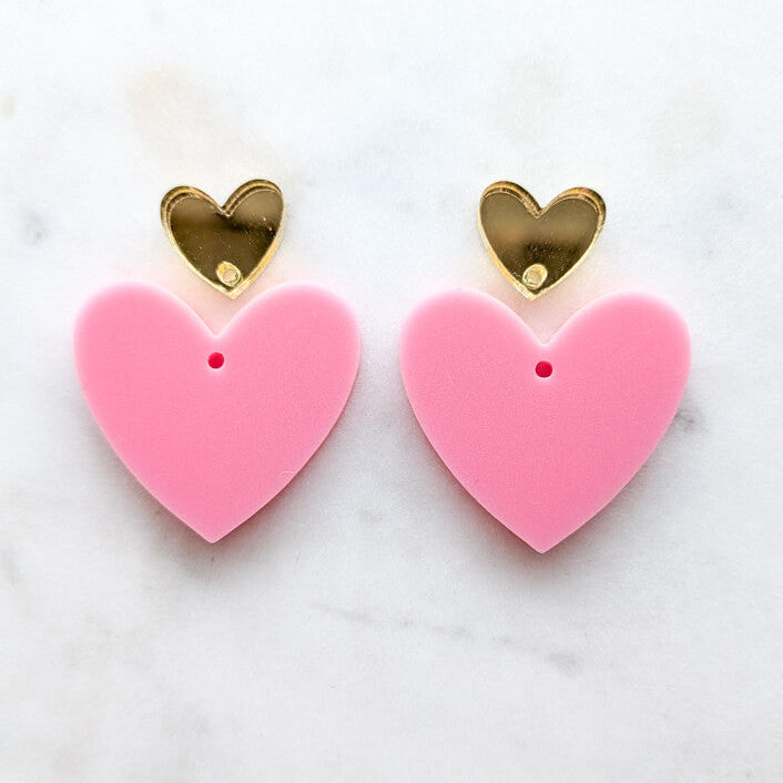 A pair of heart-shaped stud earrings with large pink puffy hearts and smaller shiny gold heart studs on a white marble background.