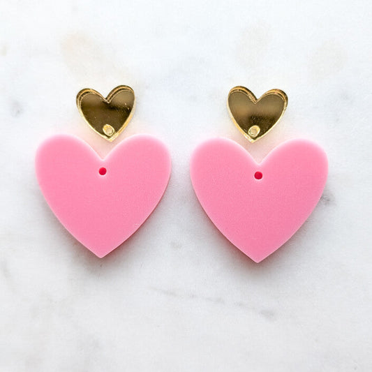 A pair of heart-shaped stud earrings with large pink puffy hearts and smaller shiny gold heart studs on a white marble background.